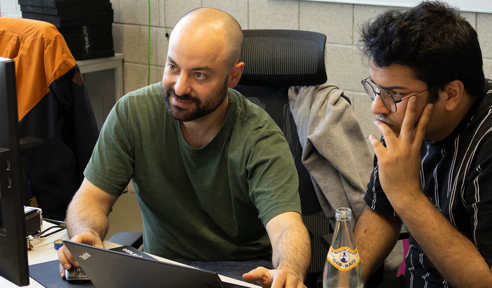 two men sitting at a desk looking at a laptop