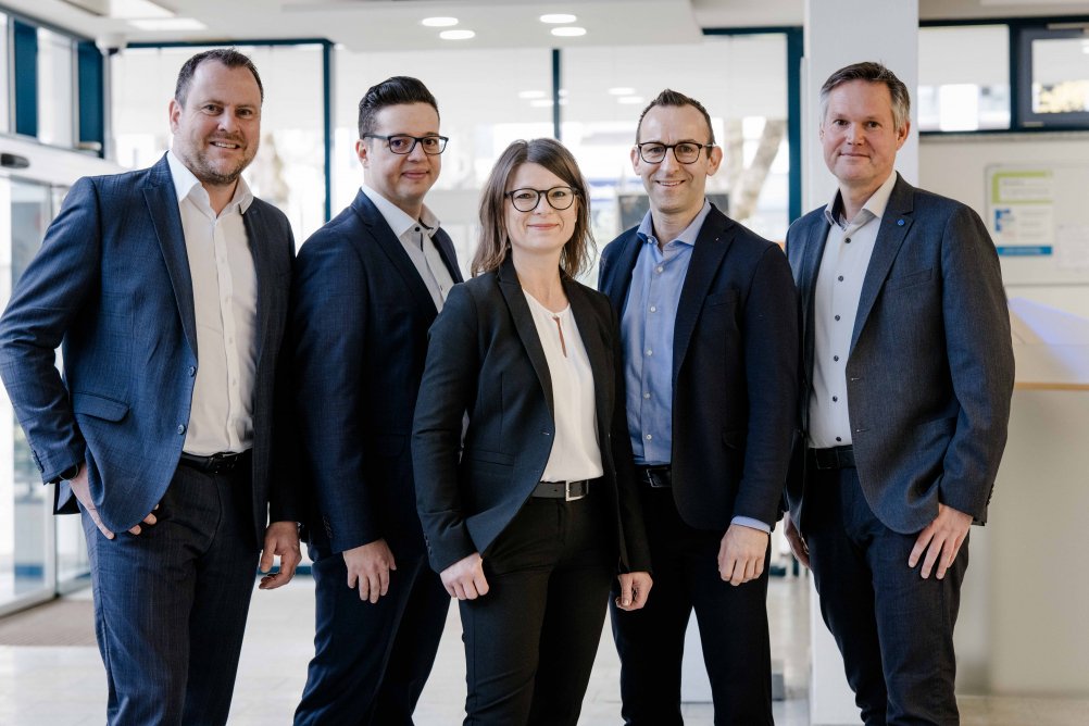 Fünf Mitarbeiter einer Genossenschaftsbank stehen gemeinsam in moderner Büroumgebung
