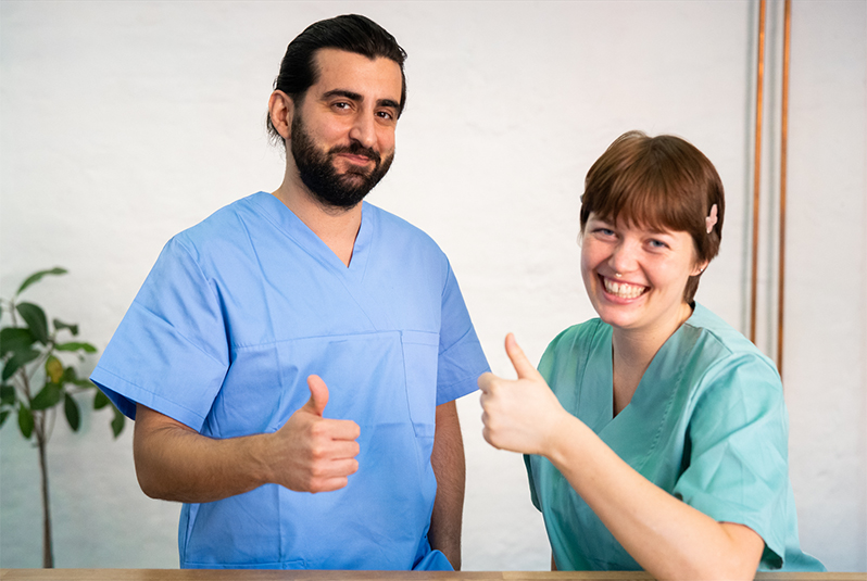 Zwei Pflegekräfte in Kasacks lächeln und geben einen Daumen hoch