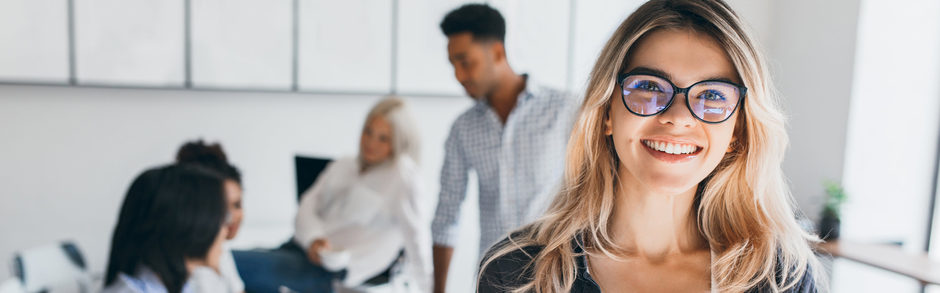 Lächelnde junge Frau mit Brille im modernen Büro mit Team im Hintergrund