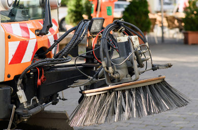 Straßenreinigungsmaschine mit orangem Fahrzeug und Kehrwalze auf Pflaster