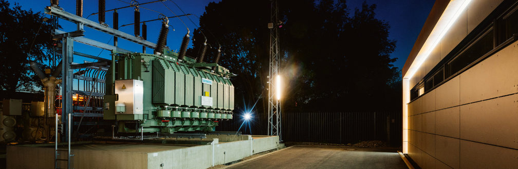 Grüner Stromtransformator in Industrieanlage bei Nacht, beleuchtet mit Hochspannungsleitungen