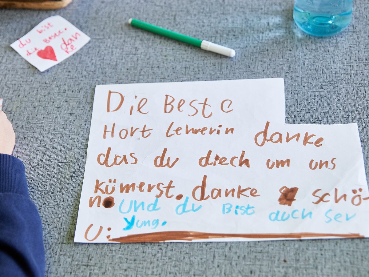 Eine Kinderhand schreibt zwei handgeschriebene Dankesbriefe an eine Lehrerin. Ein Zettel liest "Die Beste Hort Lehrerin, danke.".