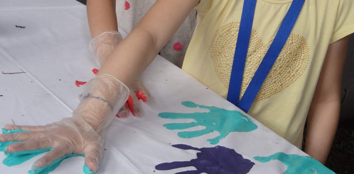 Kinder mit Plastikhandschuhen machen Handabdrücke in türkiser, violetter und roter Farbe auf einem weißen Tisch