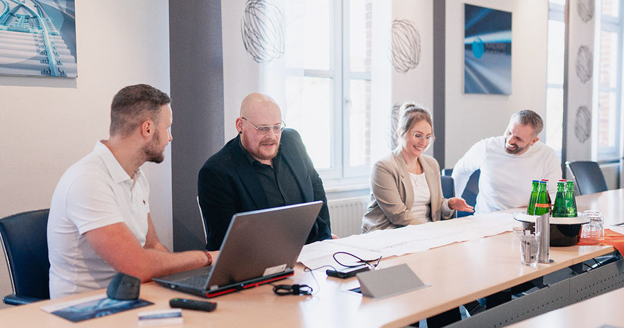 Vier Kollegen besprechen in einem Konferenzraum an einem Laptop und einem großformatigen Plan