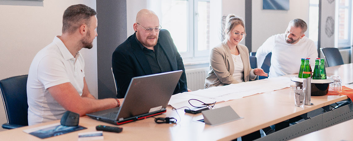 Vier Personen besprechen gemeinsam an einem Konferenztisch Pläne, vor ihnen ein aufgeklappter Laptop und Wasserflaschen