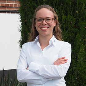 Ann-Kathrin Hippedinger, lächelnd, mit Brille, verschränkten Armen vor einer Hecke