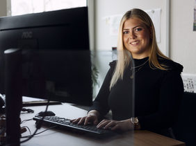 Lächelnde junge Frau mit langen blonden Haaren arbeitet am Computer in einem Büro