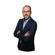 Porträt eines Mannes mit Brille und dunklem Anzug, die Arme verschränkt, vor weißem Hintergrund