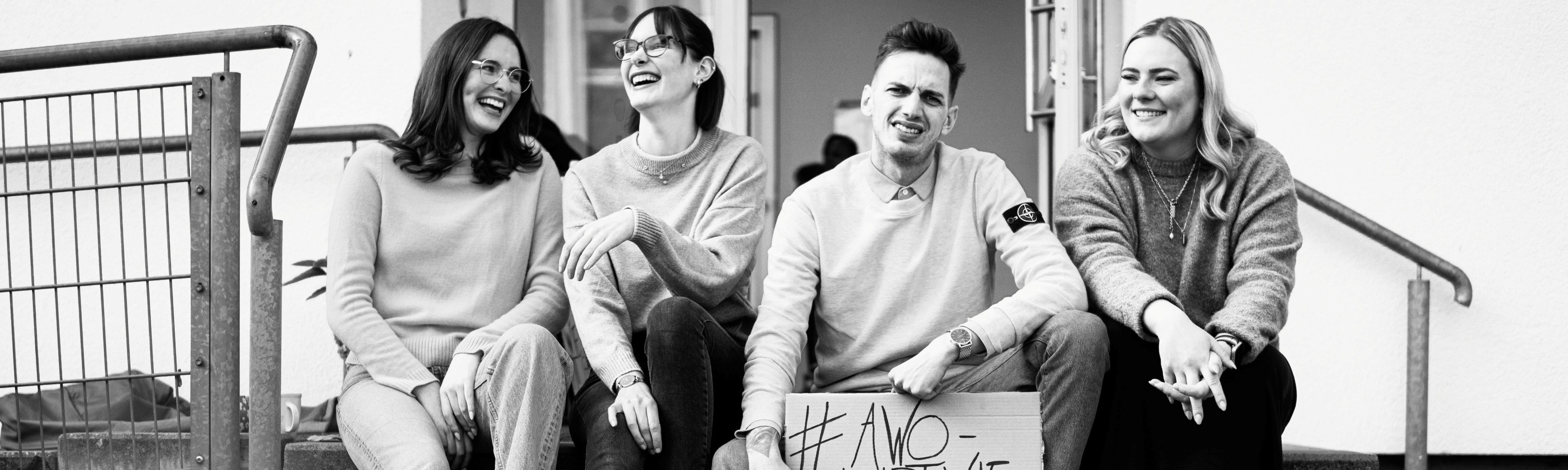 Vier lachende Teammitglieder sitzen auf einer Treppe, einer hält ein Schild mit #AWO-Hashtag