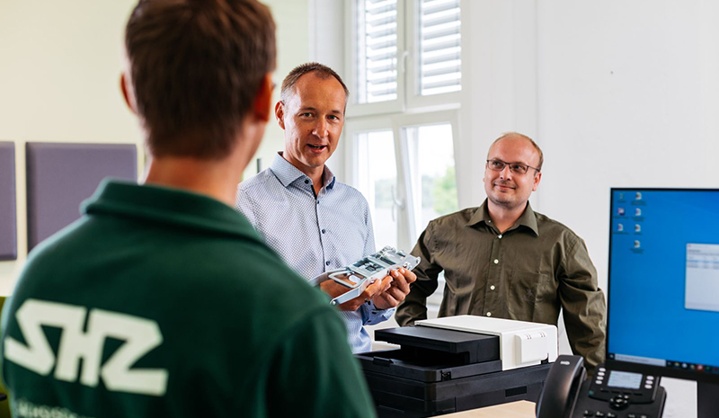 Drei Mitarbeiter  begutachten gemeinsam ein technisches Bauteil im Büro