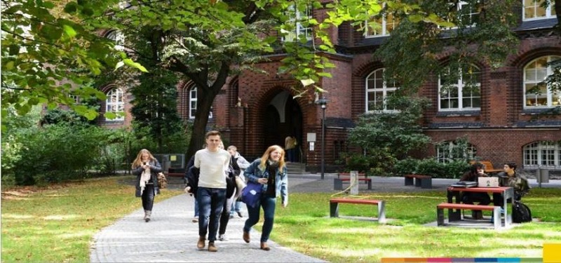 Studenten vor einem historischen Backsteingebäude auf einem grünen Campus mit Bäumen und Bänken