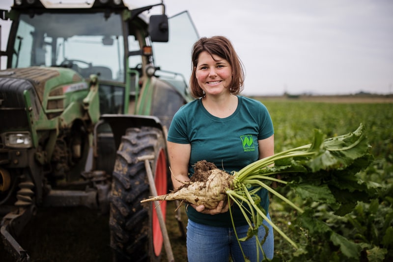 Lächelnde Frau hält Zuckerrübe auf einem Feld vor einem Traktor