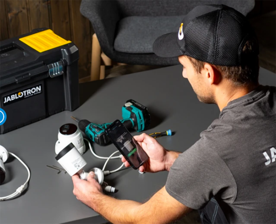 Jablotron-Techniker richtet Überwachungskameras mit einem Smartphone ein, neben einem Werkzeugkoffer und einem Akkubohrer auf einem grauen Tisch